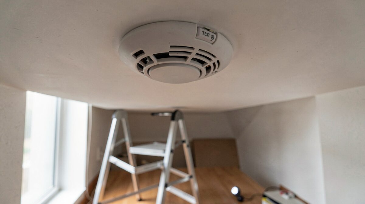 A hallway ceiling smoke alarm seen from below, its test button and vent slots in focus, a step stool and a flashlight on the floor beneath it, morning light from a nearby window.